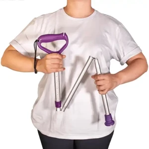 Foto centralizada em fundo branco de uma pessoa vestindo camiseta branca e calça preta, segurando de forma articulada uma bengala dobrável de alumínio prata. A mão direita segura a seção superior da bengala pelo cabo ergonômico de plástico roxo, que possui uma alça de punho preta cordada. A estrutura metálica da bengala desce dividida em três gomos conectados por uma corda elástica interna oculta, formando um desenho em zigue-zague. A mão esquerda segura a última haste inferior do bastão próximo à ponteira de borracha roxa antiderrapante com ranhuras em sua base. Na haste superior móvel, observam-se furos sequenciais para regulagem de altura por pino de pressão.