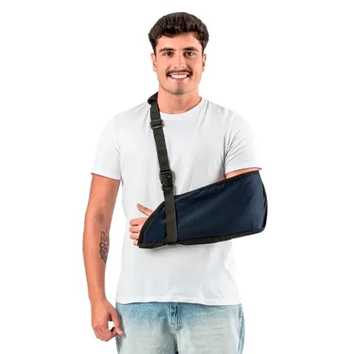 Foto em plano médio sob fundo branco isolado exibindo um homem sorrindo de cabelo escuro e bigode, vestindo uma camiseta branca lisa e calça jeans clara, enquanto utiliza o modelo Tipoia Brim Basic.webp no braço esquerdo. O suporte ortopédico de tecido de brim azul-escuro envolve confortavelmente o antebraço e o cotovelo, apresentando acabamento em costura com viés preto por toda a borda da bolsa protetora. O dispositivo é sustentado por uma alça de fita trançada preta que cruza o tórax diagonalmente em direção ao ombro direito, sendo equipada com uma fivela plástica de engate rápido e um passador retangular preto para regulagem fina de altura, mantendo o punho e a mão bem posicionados.
