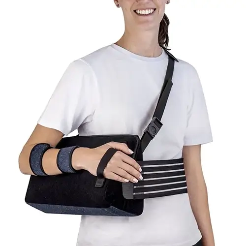 Foto em plano médio sob fundo branco isolado exibindo uma mulher sorrindo de camiseta branca que veste o modelo Tipoia de Abdução.webp no braço direito. O equipamento ortopédico preto e azul-marinho destaca um grande coxim de abdução de poliuretano posicionado lateralmente junto à cintura. O antebraço da paciente repousa sobre a almofada, fixado firmemente por duas tiras acolchoadas em azul-marinho mesclado com bordas pretas. Uma tira preta fina envolve a palma da mão direita para descanso e posicionamento ergonômico. O sistema de suspensão é composto por uma alça ajustável preta com fivela de engate rápido que cruza o tórax em direção ao ombro esquerdo e uma cinta abdominal elástica preta com faixas horizontais cinzas que envolve a região lombar para total imobilização.