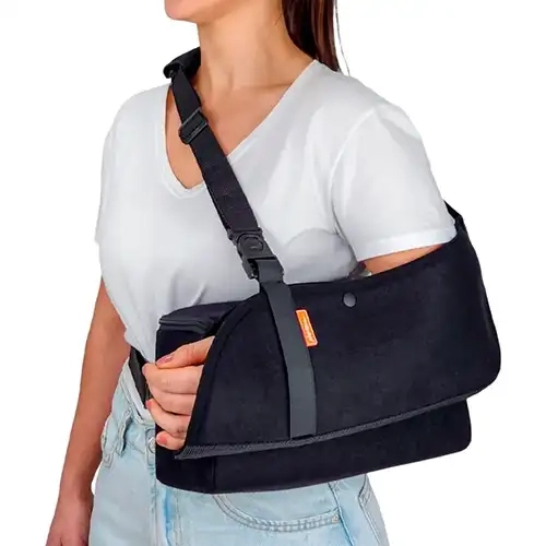 Foto em plano médio recortada sob fundo branco isolado exibindo o perfil lateral de uma mulher vestindo uma blusa de gola V branca e calça jeans clara enquanto utiliza o modelo Tipoia de Abdução_2.webp no braço esquerdo. O dispositivo ortopédico na cor preta apresenta uma bolsa de suporte aveludada para o antebraço apoiada perfeitamente sobre um coxim de abdução retangular espesso de espuma densa preta posicionado junto à cintura. A tipoia conta com um botão de pressão frontal discreto, uma pequena etiqueta retangular laranja com detalhes em branco costurada na borda superior e uma fita de apoio vertical preta que segura a região do punho e da mão. O sistema de suspensão é composto por uma alça de fita trançada preta ajustável com fivela de engate rápido e almofada protetora estofada sobre o ombro direito, além de uma tira estabilizadora preta que contorna a região lombar prendendo o coxim firmemente ao tronco.