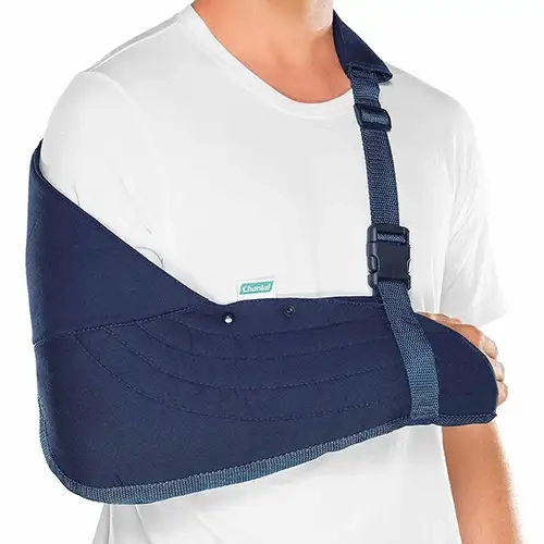 O dispositivo ortopédico na cor azul-marinho envolve completamente o cotovelo e o antebraço em uma bolsa de sarja acolchoada com costuras horizontais curvas e acabamento de viés cinza nas bordas. Uma aba de tecido estende-se para cima cobrindo a articulação do ombro direito, trazendo uma pequena etiqueta branca e verde da marca Chantal fixada na borda superior, com dois botões de pressão pretos logo abaixo. O sistema de suspensão conta com uma alça de tecido trançado azul-marinho com almofada protetora estofada sobre o ombro esquerdo, equipada com passadores de regulagem pretos e uma fivela de engate rápido de poliamida preta conectada verticalmente à base do antebraço.
