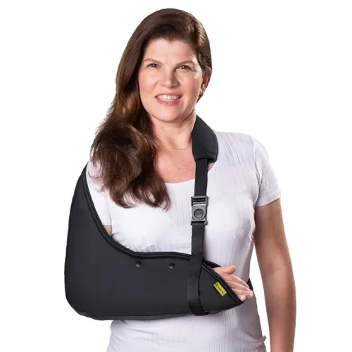 Foto em plano médio sob fundo branco isolado exibindo uma mulher de cabelos castanhos longos e blusa branca que veste o modelo Tipoia Velpeau Estofada.webp no braço direito. O suporte ortopédico na cor preta envolve completamente o cotovelo e o antebraço em uma bolsa de tecido lisa e acolchoada de poliuretano, com dois ilhoses de respiro pretos na lateral e uma pequena etiqueta retangular amarela da marca Kestal costurada na extremidade frontal próxima à mão. O sistema de suspensão conta com uma alça regulável preta com almofada protetora estofada sobre o ombro esquerdo, equipada com uma fivela de encaixe e regulador de poliamida preta conectada verticalmente para sustentar o punho. Uma faixa estabilizadora preta estende-se horizontalmente por trás da cintura para fixação completa do membro.