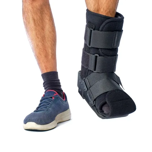 Foto em plano médio mostrando as pernas de uma pessoa; o pé direito está utilizando uma bota imobilizadora ortopédica preta de cano longo com tiras de velcro, enquanto o pé esquerdo calça um tênis esportivo azul com detalhes vermelhos e meia escura, sobre um fundo branco.