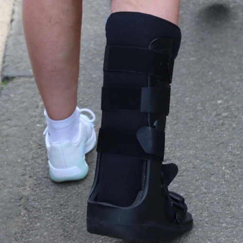 Vista posterior das pernas de uma pessoa caminhando; o pé direito utiliza uma bota imobilizadora ortopédica preta de cano longo e o pé esquerdo um tênis esportivo branco com meia branca, sobre solo asfáltico.