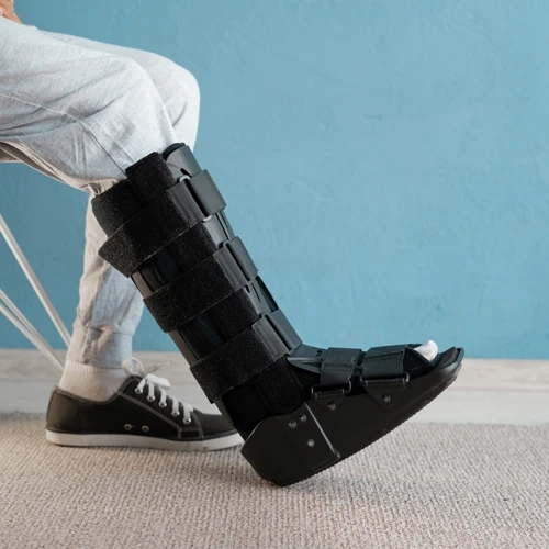 Paciente sentado utilizando uma bota imobilizadora ortopédica preta de cano longo com múltiplos fechos de velcro, vista de perfil contra uma parede azul claro e piso cinza.